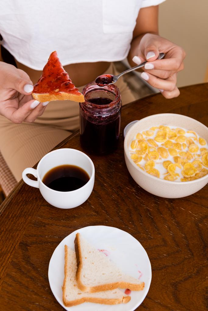 vista cortada da mulher americana africana segurando colher com geléia perto de pão torrado, flocos de milho na tigela e xícara com café na mesa
  - Foto, Imagem