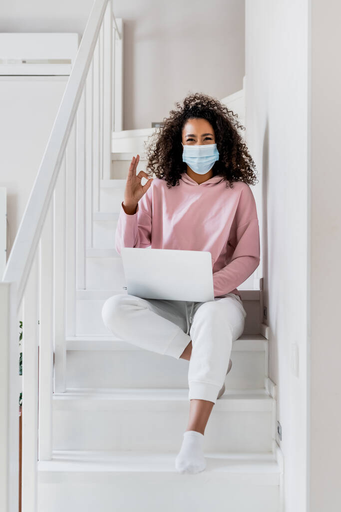 rizado afroamericano freelancer en máscara médica sentado en escaleras y mostrando ok signo cerca de la computadora portátil
  - Foto, Imagen