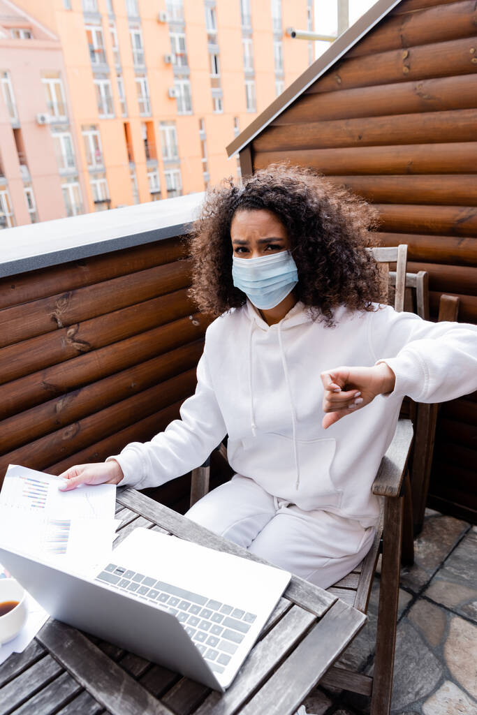 chica afroamericana en máscara médica utilizando portátil cerca de gráficos y gráficos
  - Foto, Imagen