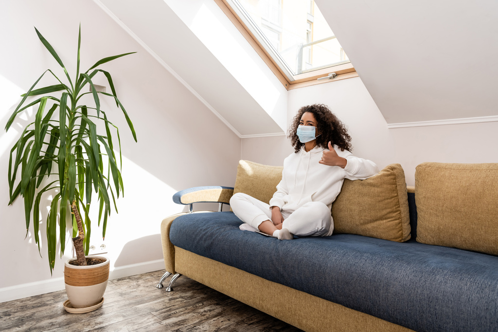chica americana africana rizada en máscara médica mostrando el pulgar hacia arriba mientras está sentado en el sofá
  - Foto, imagen
