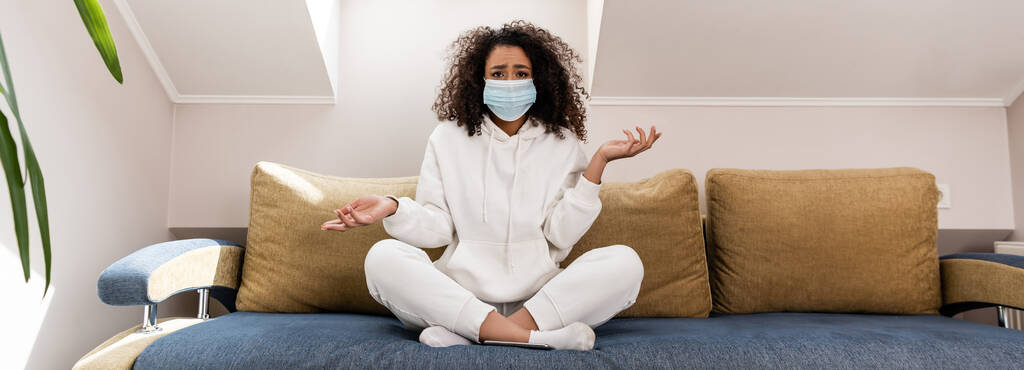οριζόντια έννοια του σγουρού αφροαμερικανού κοριτσιού σε ιατρική μάσκα gesturing, ενώ κάθεται στον καναπέ - Φωτογραφία, εικόνα
