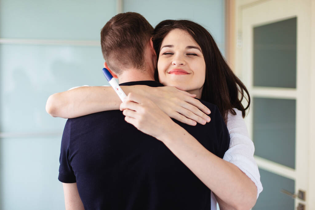Молода жінка обіймає чоловіка з позитивним тестом на вагітність в руці. Щасливі майбутні батьки чекають на дитину
 - Фото, зображення