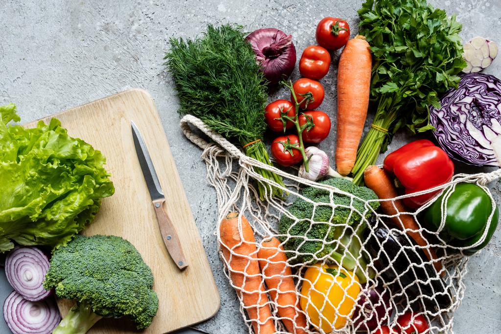 vista superior de verduras frescas maduras en bolsa de hilo cerca de la tabla de cortar con cuchillo en la superficie de hormigón gris
 - Foto, Imagen