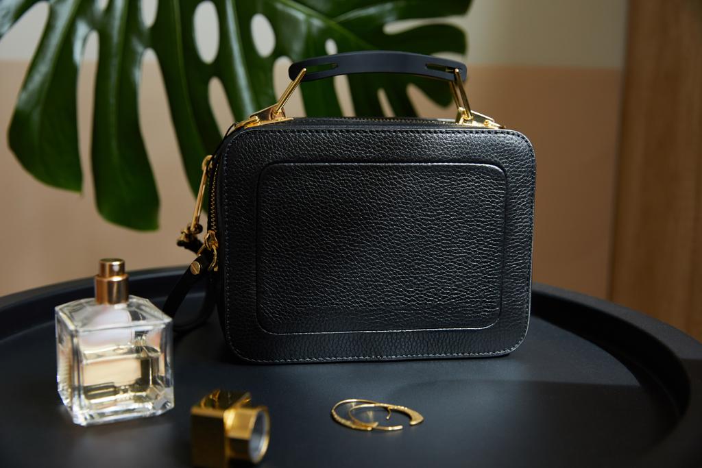 bolso de cuero cerca de pendientes de oro, perfume en mesa negra cerca de hoja tropical
 - Foto, imagen