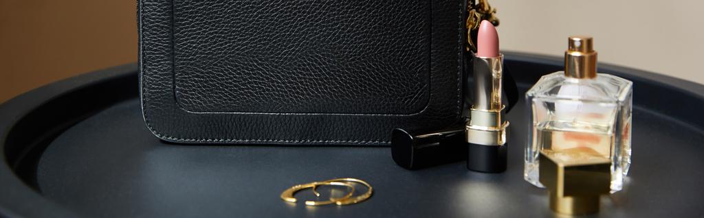 leather handbag near golden earrings, perfume and lipstick on black table, panoramic shot - Photo, Image