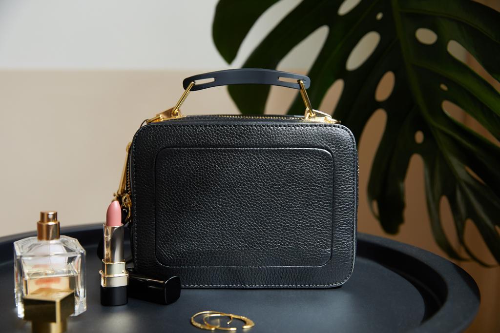kožená kabelka poblíž zlatých náušnic, parfémů a rtěnky na černém stole poblíž tropického listu - Fotografie, Obrázek