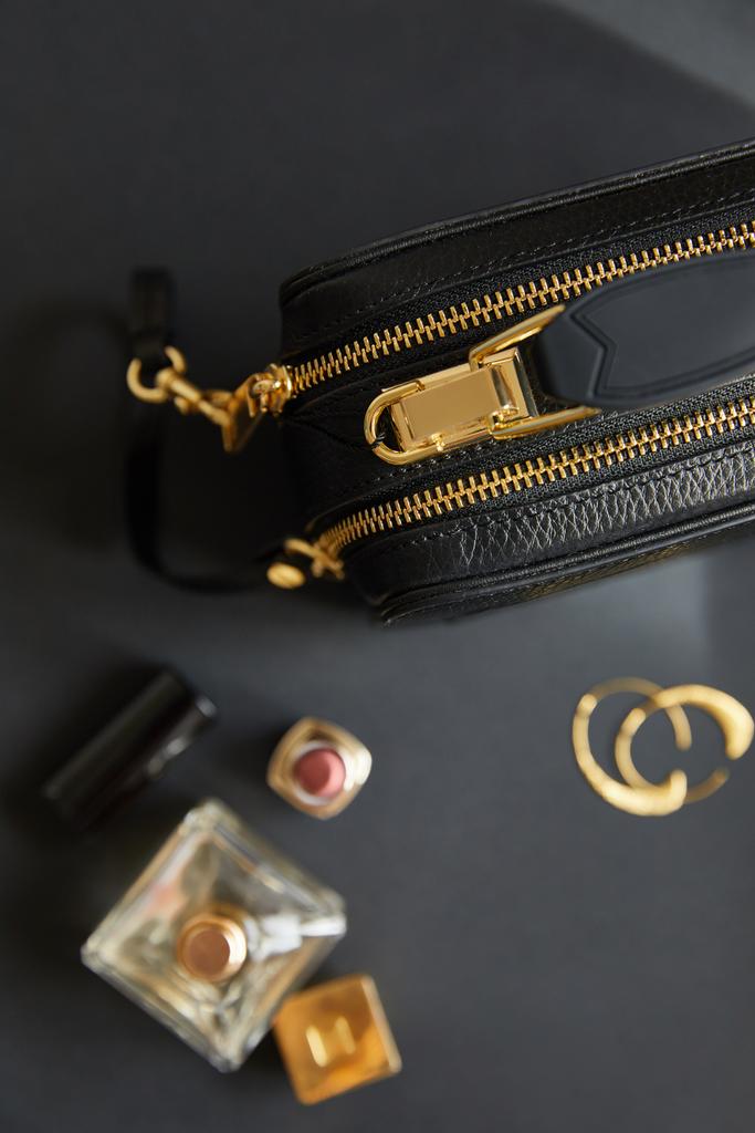 Ansicht von Lederhandtasche in der Nähe goldener Ohrringe, Parfüm und Lippenstift auf schwarzem Tisch - Foto, Bild