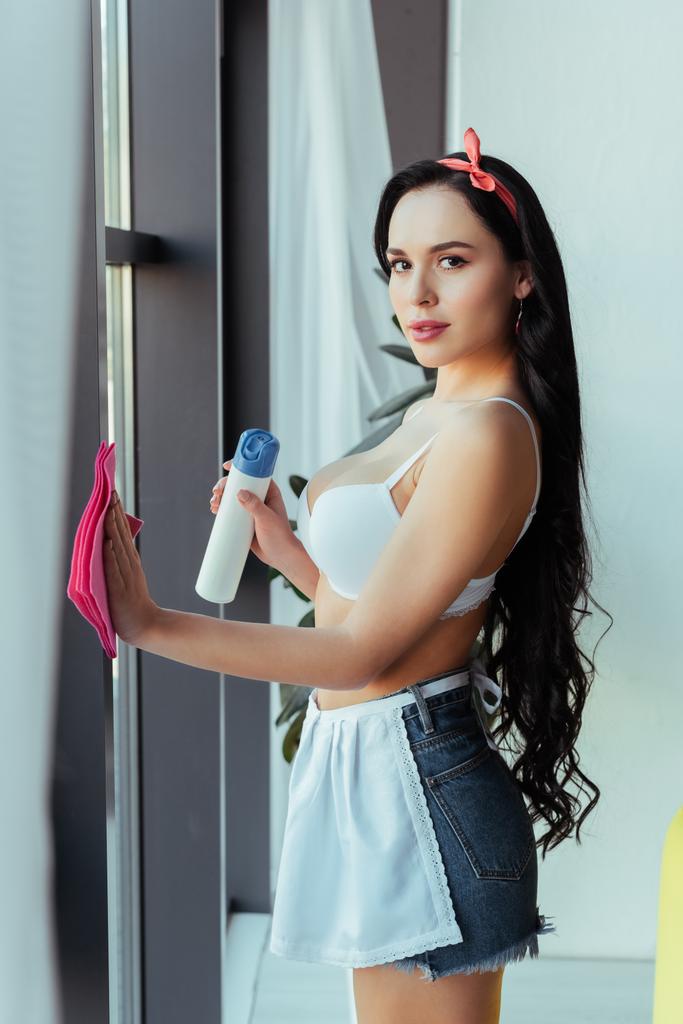 Widok z boku zmysłowy dziewczyna w biustonosz i fartuch czyszczenia okna z szmatą w domu  - Zdjęcie, obraz