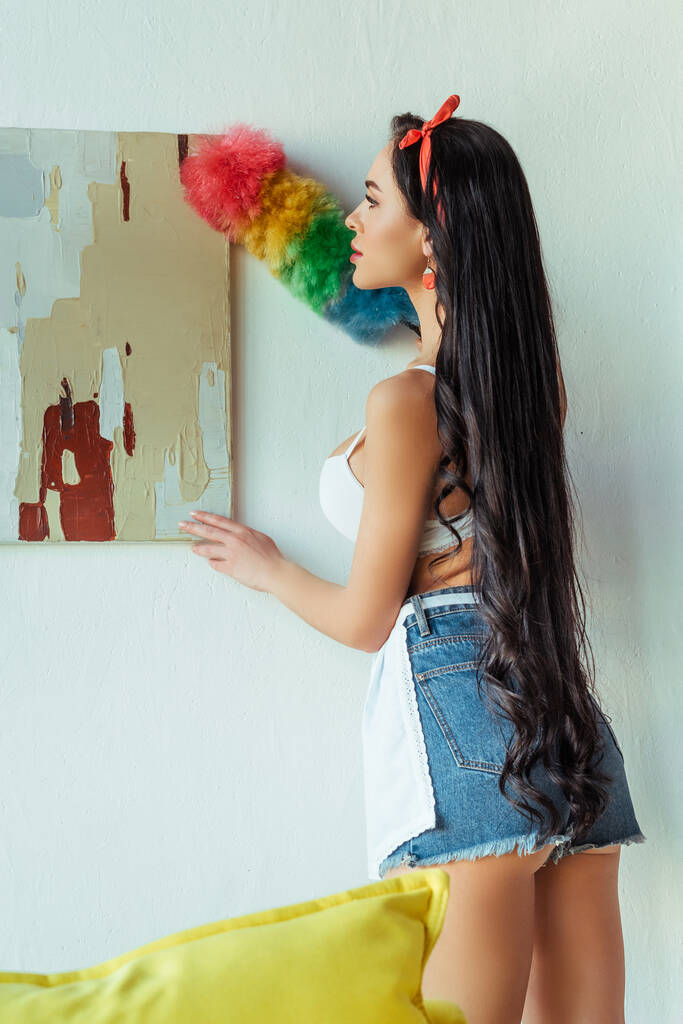Vue arrière de la femme sensuelle en soutien-gorge et tablier nettoyage peinture avec brosse à poussière
  - Photo, image