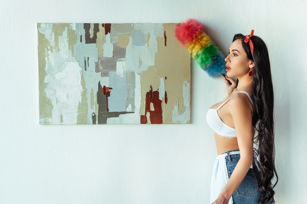 Seitenansicht von sexy Mädchen in BH und Schürze Putzen Malerei mit Staubpinsel zu Hause  - Foto, Bild