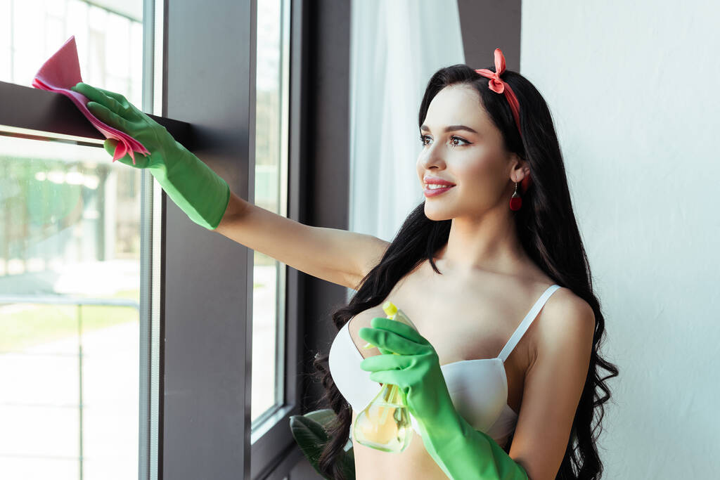 Чуттєва дівчина посміхається при чищенні вікна з ганчіркою і пляшкою розпилення
 - Фото, зображення