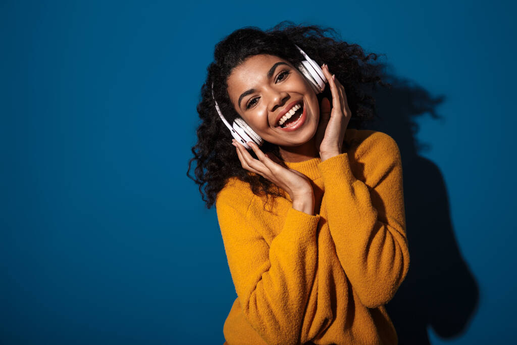 Foto de feliz positivo jovem africana posando isolado sobre fundo parede azul ouvindo música com fones de ouvido
. - Foto, Imagem