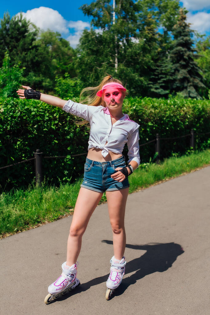 Portret emocjonalnej pięknej młodej dziewczynki w różowym daszkiem i rękawice ochronne do rolek i skateboardingu na rolkach na drogach. - Zdjęcie, obraz