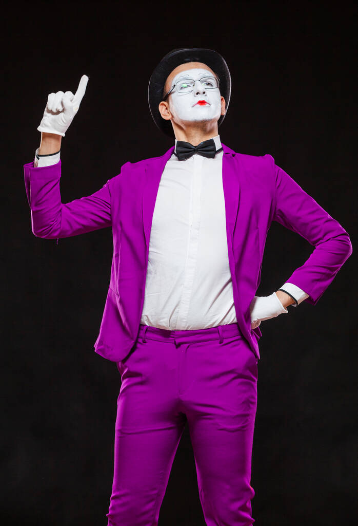 Retrato de mimo masculino, aislado sobre fondo negro. Un joven con traje púrpura está de pie con el dedo levantado. Símbolo de atención, advertencia, nota bene - Foto, imagen