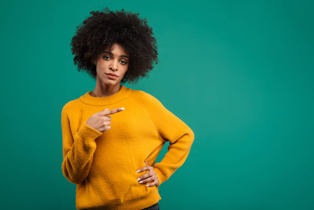 Porträt einer selbstbewussten jungen Afrikanerin im Pullover, die isoliert vor grünem Hintergrund steht und mit dem Finger auf Kopierraum zeigt - Foto, Bild
