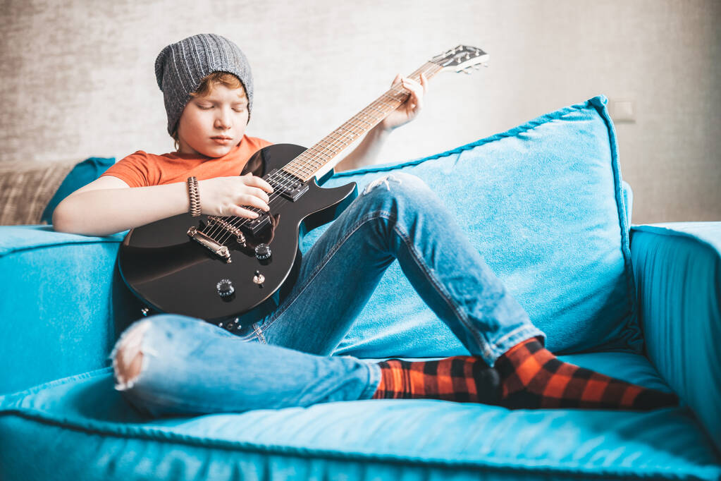Κομψό rock and roll παιδί με μαύρη ηλεκτρική κιθάρα κάθεται σε μια καρέκλα και παίζει μουσική - Φωτογραφία, εικόνα