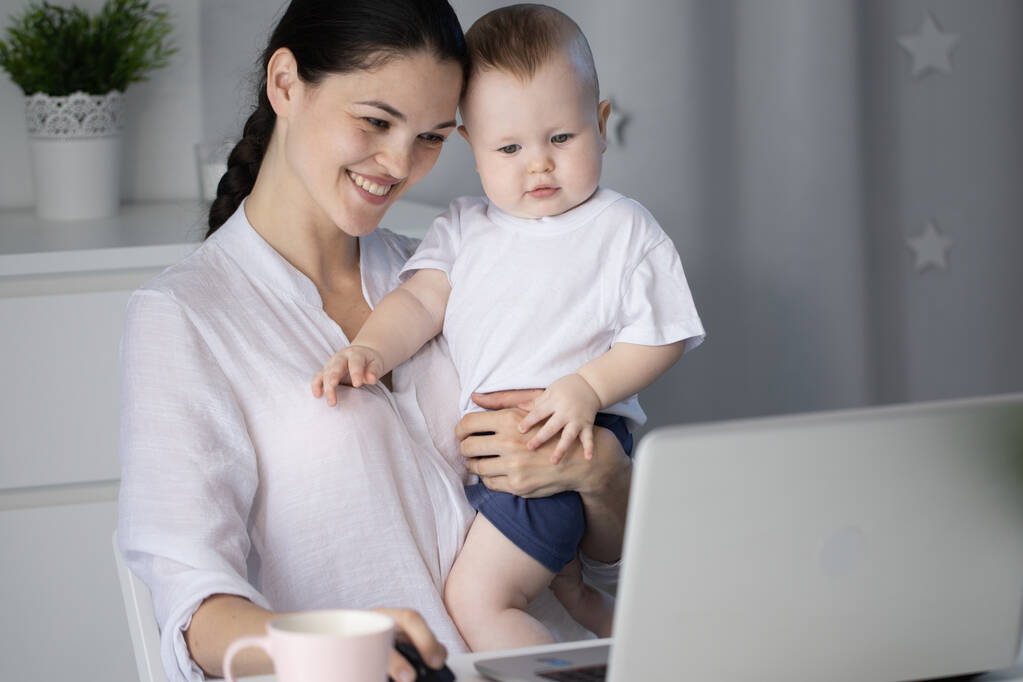 Joven madre trabajando en un decreto en la computadora con un hijo bebé en sus brazos
 - Foto, Imagen
