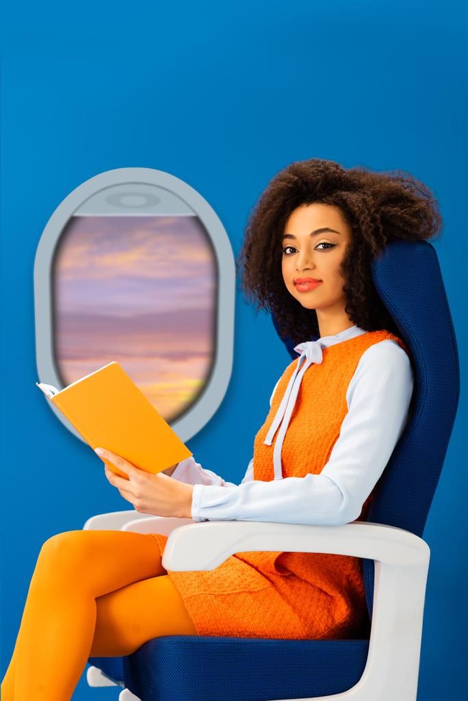 усміхнена афроамериканська жінка в ретро-сукні тримає книгу і сидить на сидінні ізольовано на синьому з отвором
 - Фото, зображення