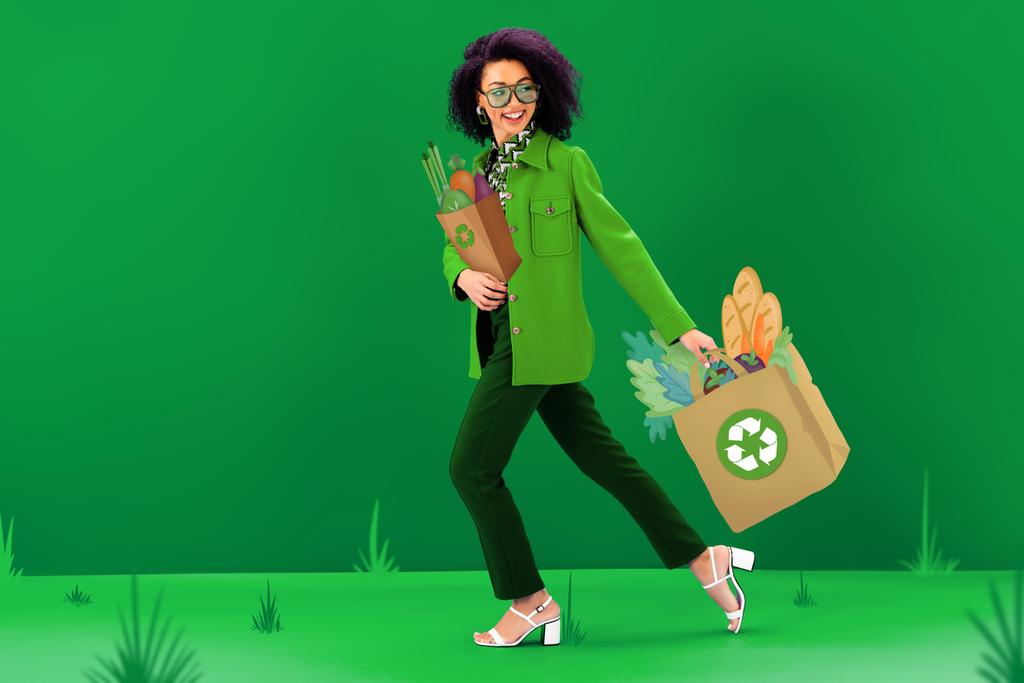 mujer afroamericana sonriente y elegante caminando con bolsas de compras ilustradas con carteles de reciclaje sobre fondo verde
  - Foto, Imagen