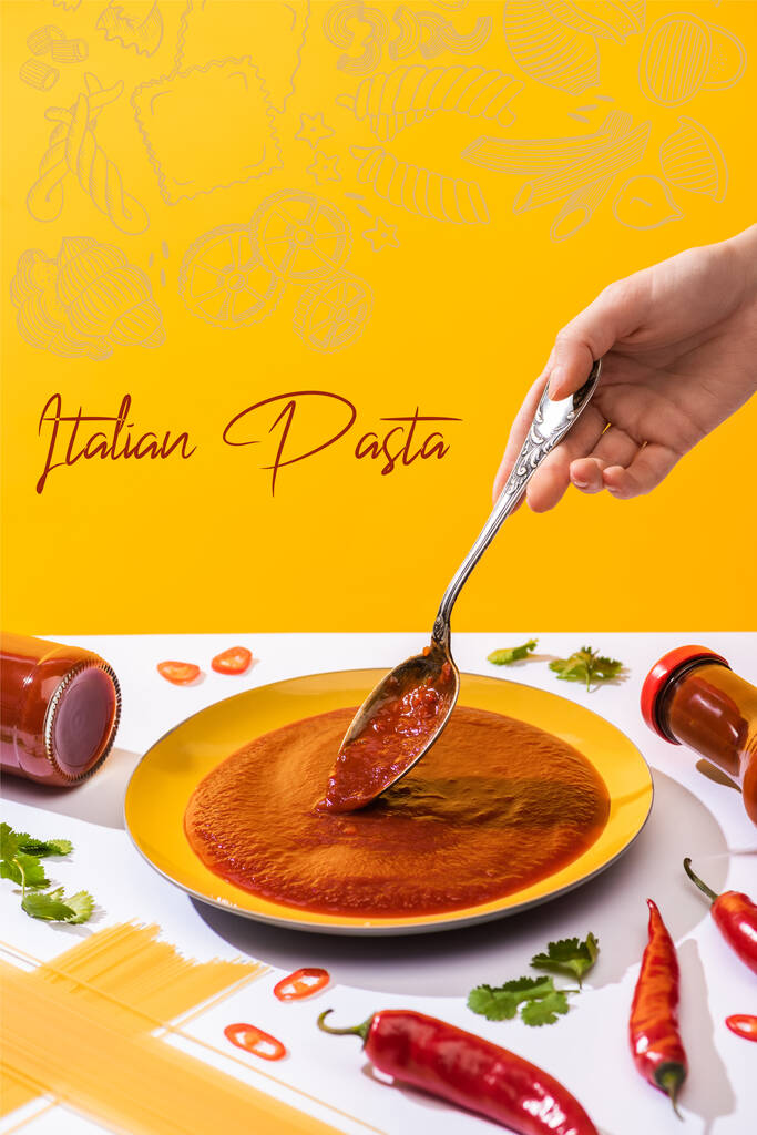 Cropped view of woman holding spoon with ketchup with spaghetti and chili peppers on white surface isolated on yellow, Italian pasta illustration - Photo, Image
