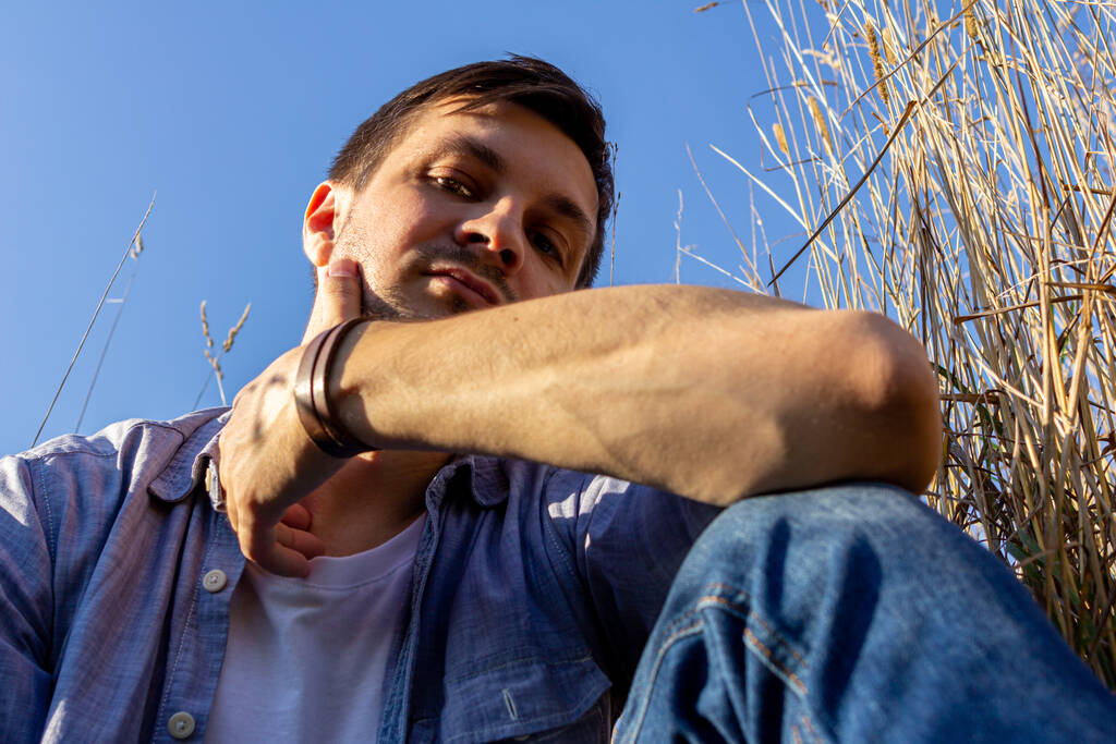 un giovane uomo attraente indossa jeans, maglietta bianca e una camicia blu si trova su un campo di cereali contro un cielo blu e guarda alla fotocamera
 - Foto, immagini