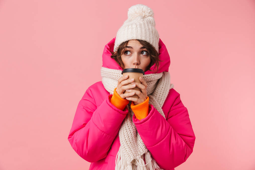 Retrato de una hermosa mujer linda en ropa de abrigo mirando a un lado y bebiendo café aislado sobre fondo rosa
 - Foto, Imagen