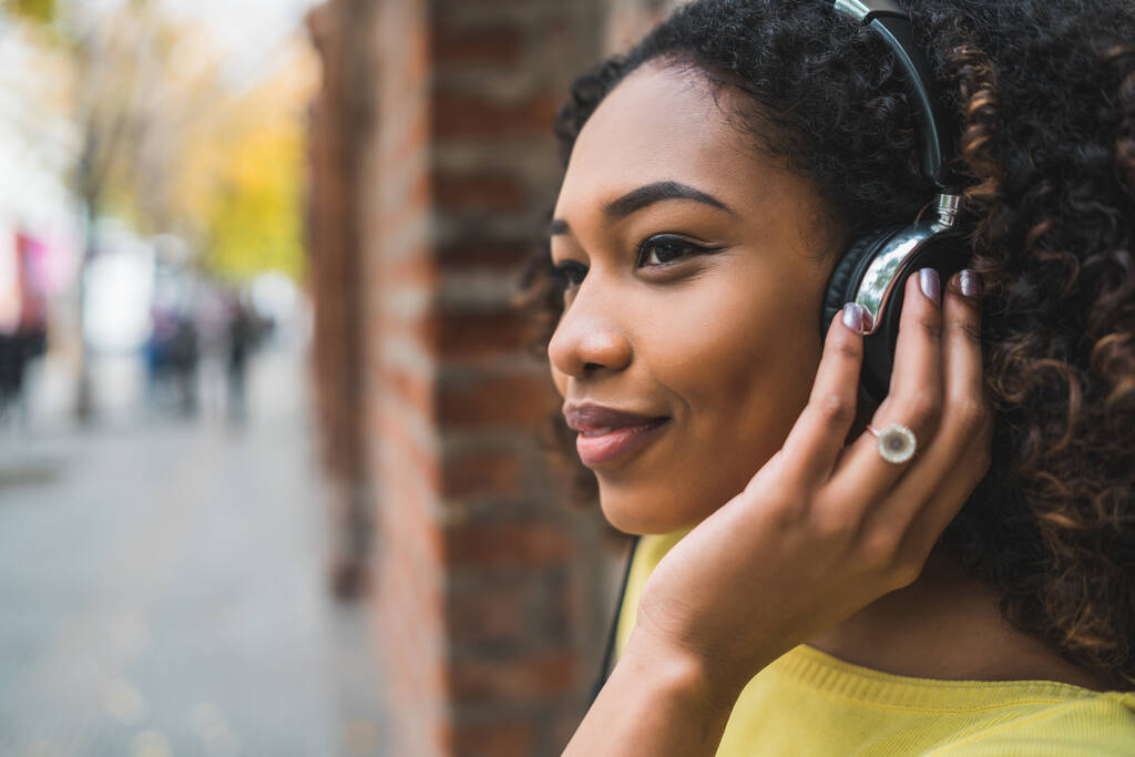 Portret młodej Afro amerykańskiej kobiety słuchającej muzyki ze słuchawkami na ulicy. Na zewnątrz. - Zdjęcie, obraz