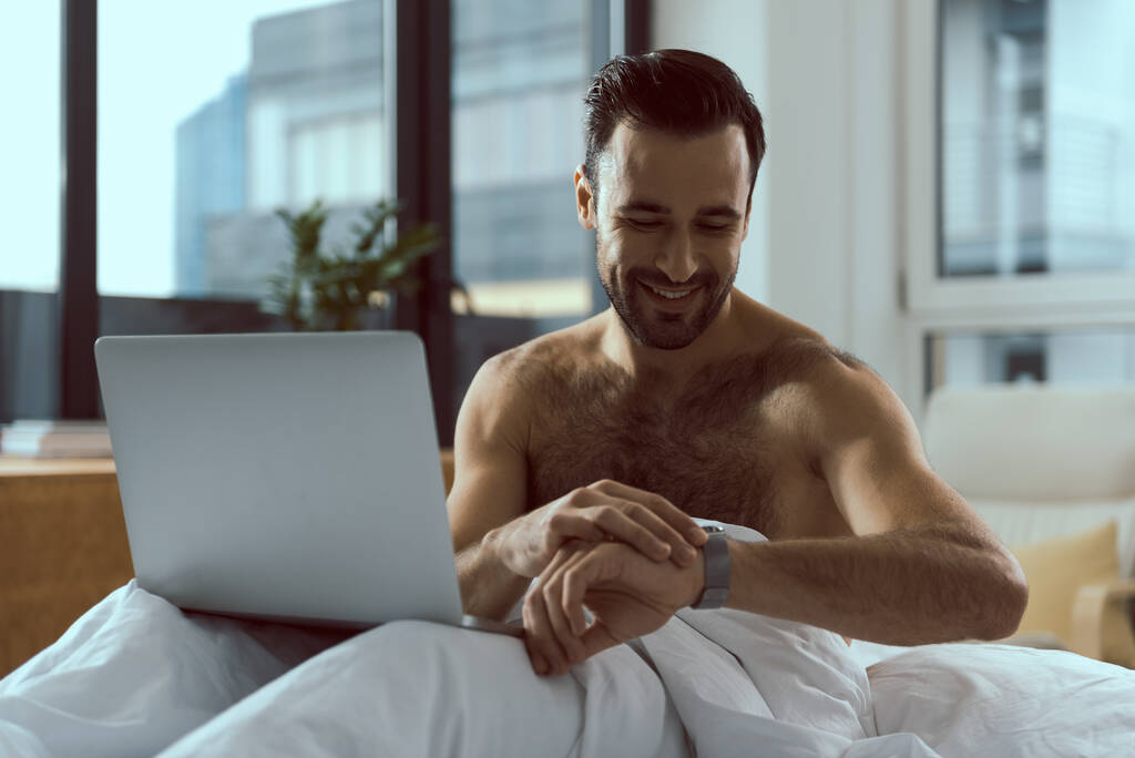 Ευτυχισμένος άνθρωπος με σημειωματάριο κοιτάζοντας ρολόι στο κρεβάτι - Φωτογραφία, εικόνα