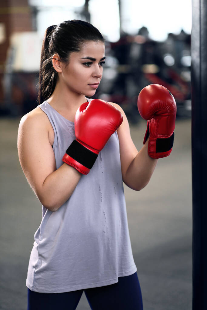 Sportliche junge brünette Frau in Sportbekleidung und roten Boxhandschuhen trainiert Beulen auf einem Boxsack in einem Fitnessstudio. - Foto, Bild
