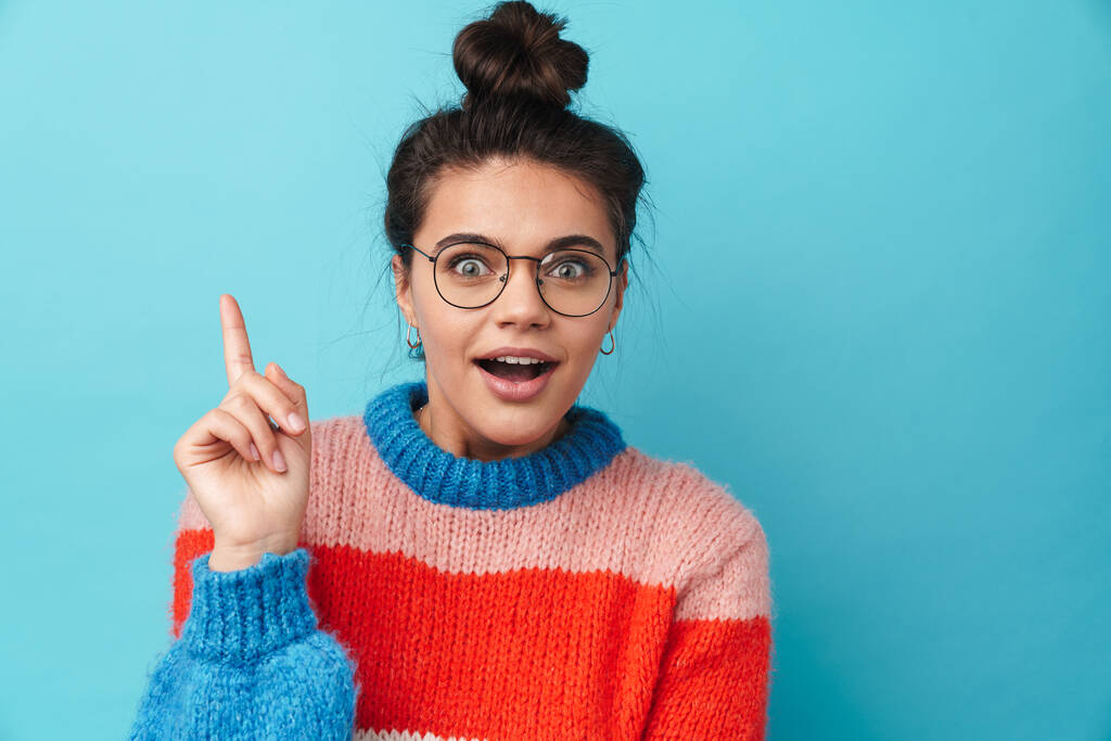 Image of excited young woman in eyeglasses expressing surprise and pointing finger upward isolated over blue background - Photo, Image