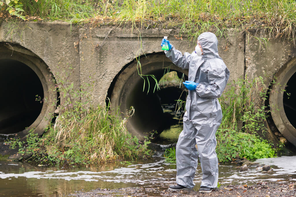 человек в защитном костюме и маске держит в руках фляжку с зеленой жидкостью на фоне канализационных труб, выходящих из завода
 - Фото, изображение