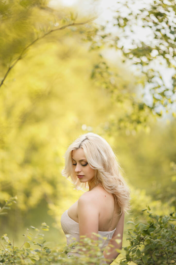 Attraktive junge blonde Frau genießt ihre Zeit draußen im Park mit grünen Bäumen im Hintergrund. - Foto, Bild