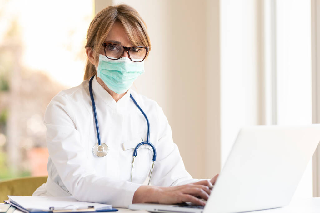Shot van overwerkte vrouwelijke arts dragen gezichtsmasker voor preventie tijdens het zitten aan het bureau en werken op laptop in het ziekenhuis. - Foto, afbeelding