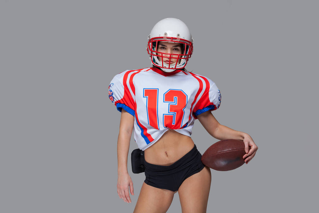 Amerikan futbolcusu ve kaskından seksi üniforma giyen sporcu kız gri arka planda topu fırlatmaya hazır. - Fotoğraf, Görsel