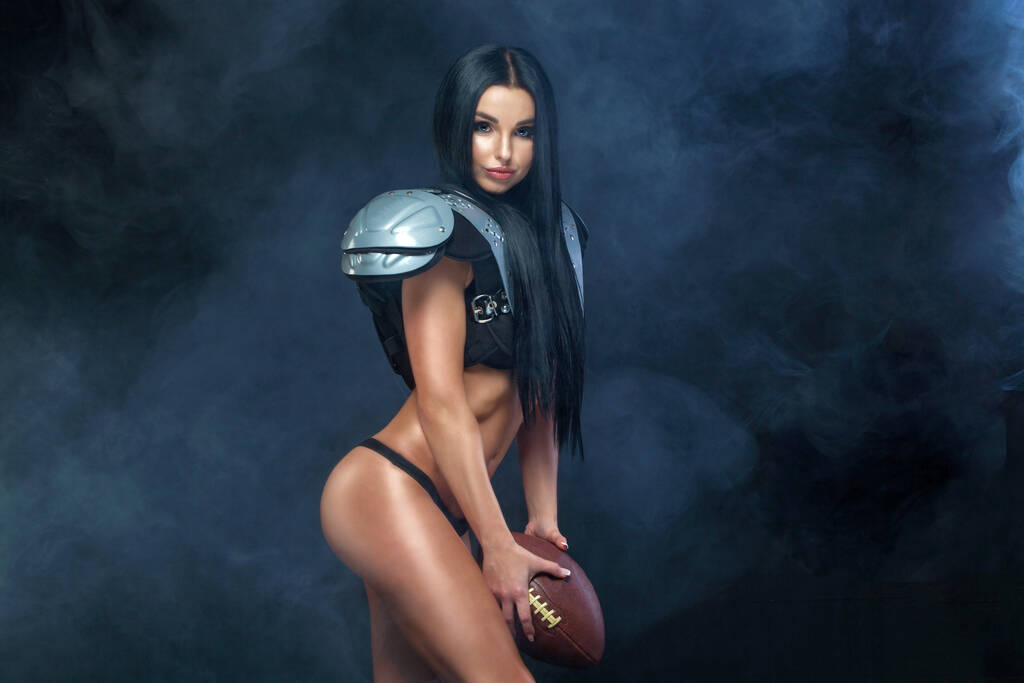 American football. Young sporty brunette wearing sexy uniform of rugby football player posing with ball in smoke isolated on black background - Photo, Image