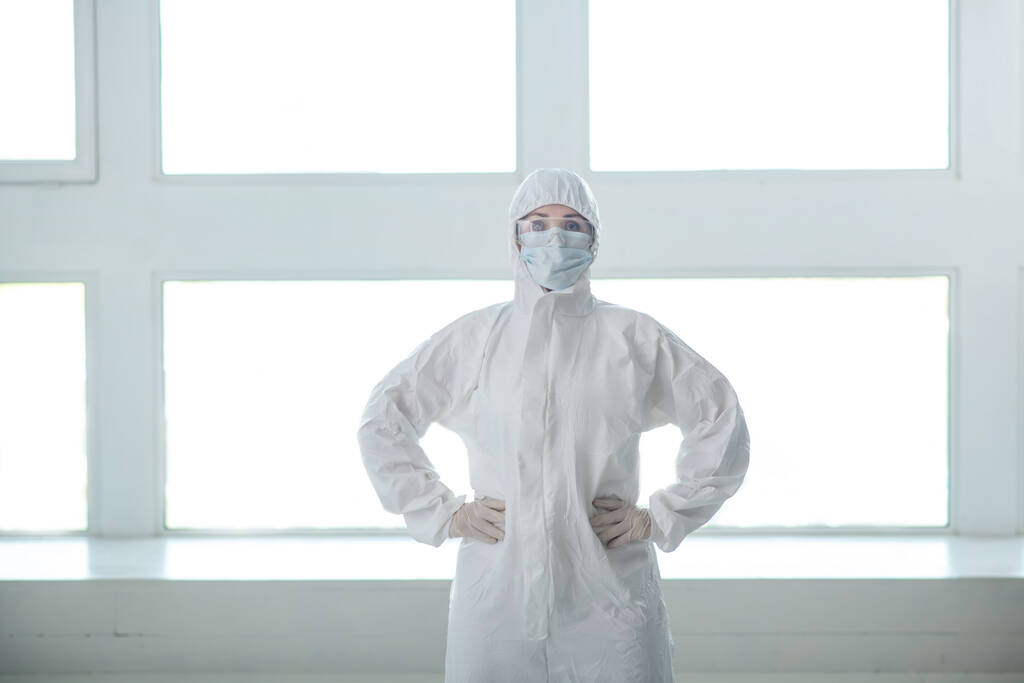 Lääketieteellinen työntekijä suojavaatteissa, lääketieteellisessä naamiossa ja suojalaseissa, jotka seisovat kädet vyötäröllä
 - Valokuva, kuva