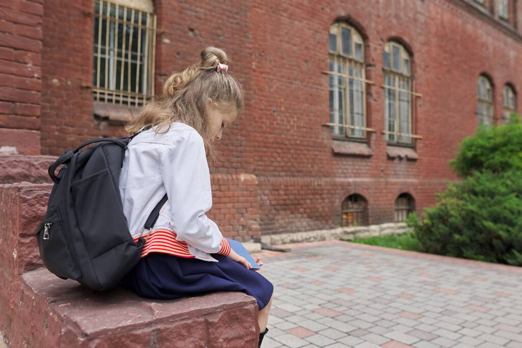 Niña con mochila sentada en el patio de la escuela leyendo cuaderno, espacio para copiar
 - Foto, imagen