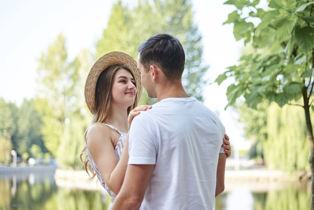 Νεαρό ζευγάρι ερωτευμένο, αγκαλιασμένο στο πάρκο το καλοκαίρι, χαμογελαστό. Όμορφο ξανθό κορίτσι σε ριγέ συνολικά και ψάθινο καπέλο σε ρομαντικό ραντεβού με όμορφο μελαχρινό άντρα σε λευκό μπλουζάκι. Κυριακή με τα πόδια. - Φωτογραφία, εικόνα
