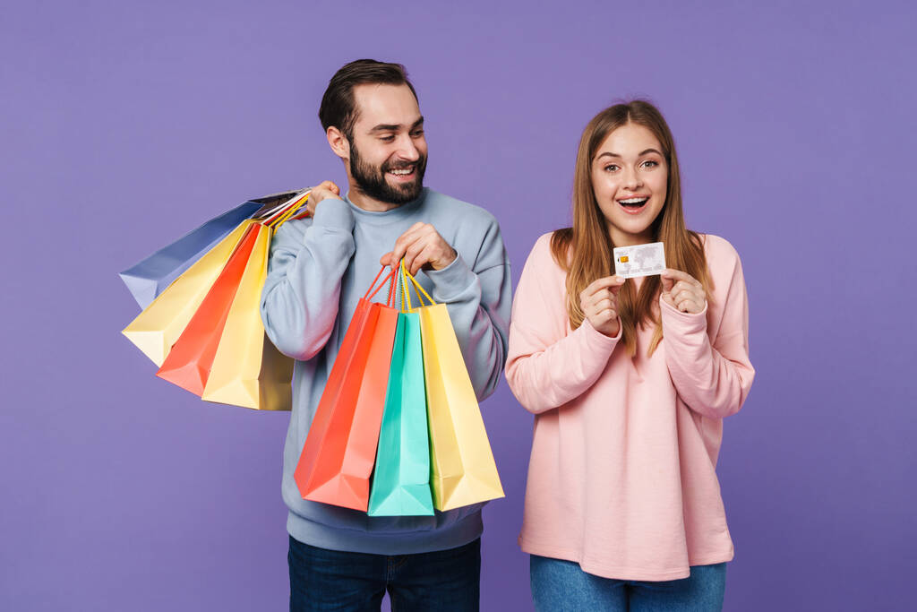 Wizerunek pozytywnej szczęśliwej kochającej się pary odizolowanej na fioletowym tle ściany trzymającej torby na zakupy i kartę kredytową. - Zdjęcie, obraz