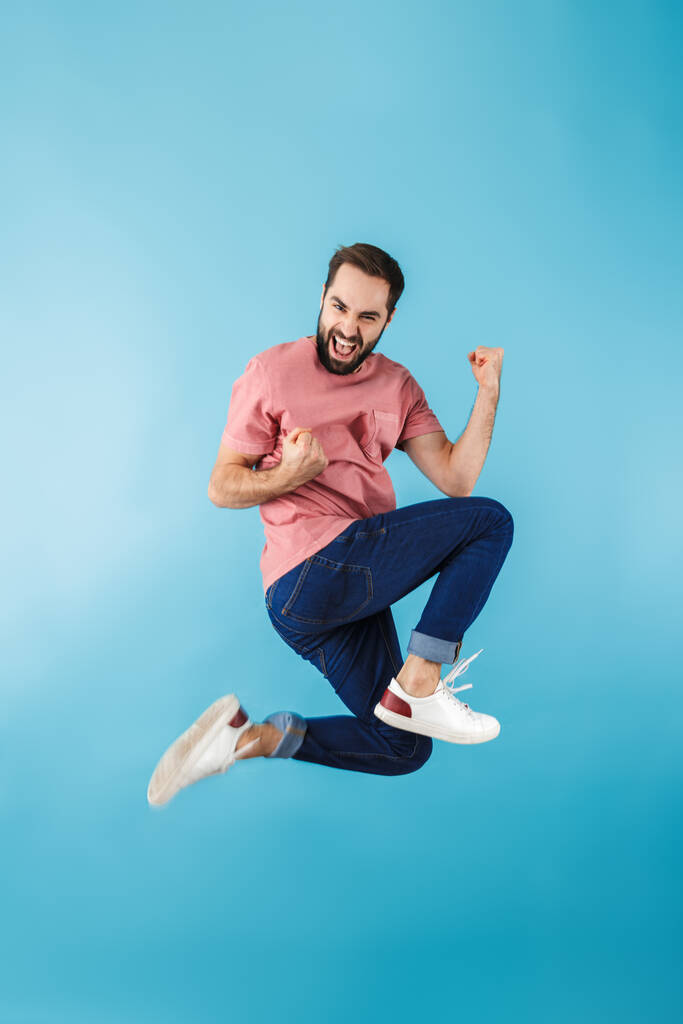 Volle Länge eines jungen, fröhlich aufgeregten, bärtigen Mannes, der ein T-Shirt trägt und isoliert über blauen Hintergrund springt - Foto, Bild