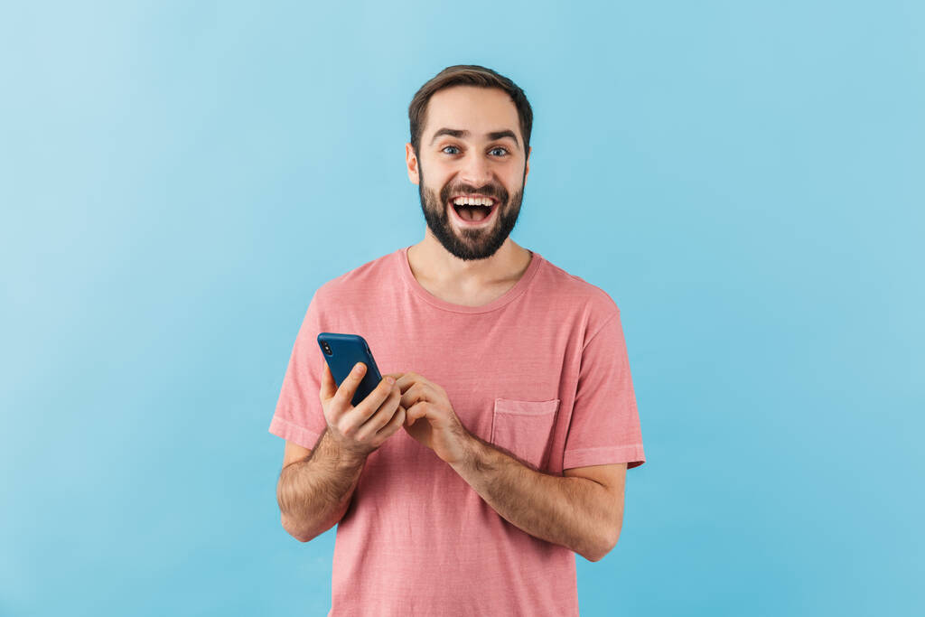 Portrait of a young cheerful excited bearded man wearing t-shirt standing isolated over blue background, using mobile phone - Photo, Image