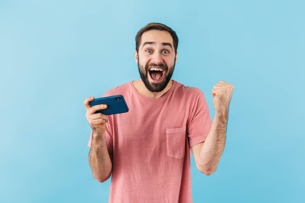 Портрет молодого веселого збудженого бородатого чоловіка в футболці, що стоїть ізольовано на синьому фоні, з використанням мобільного телефону
 - Фото, зображення