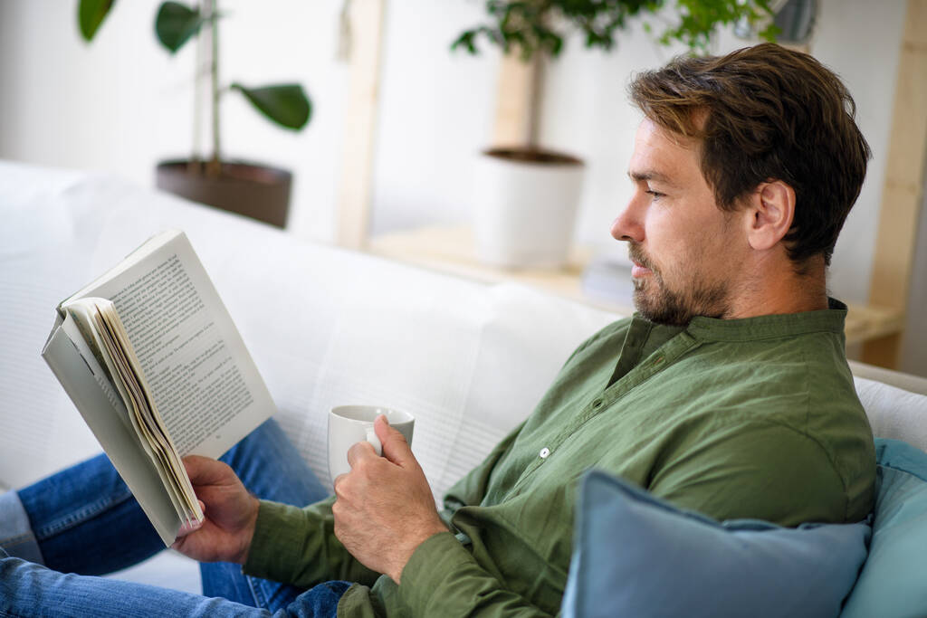 Mężczyzna siedzi na kanapie i czyta książkę w domu, koncepcja zamknięcia. - Zdjęcie, obraz