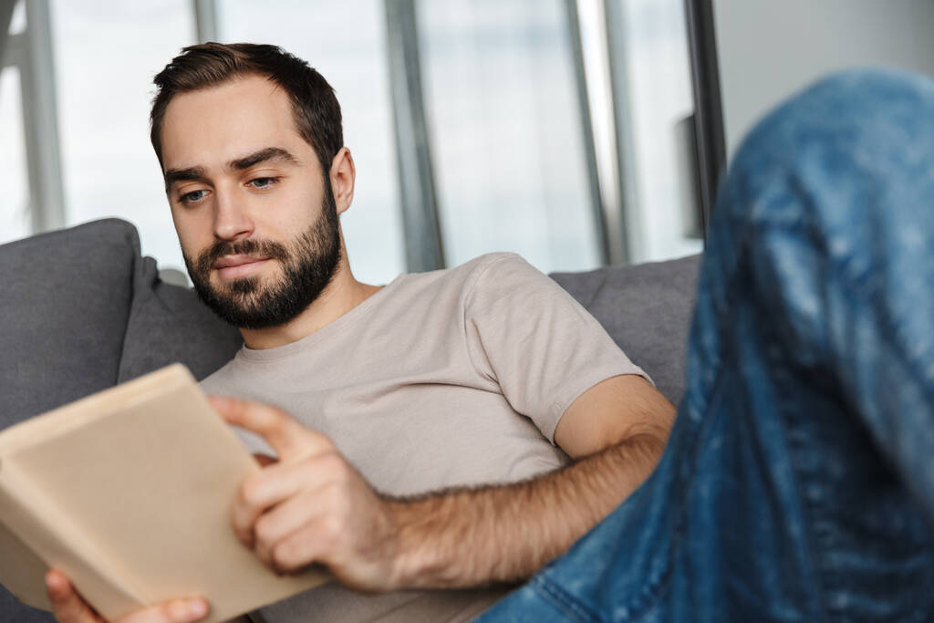 Fotografie koncentrovaného mladého pohledného muže doma na pohovce čtení knihy. - Fotografie, Obrázek