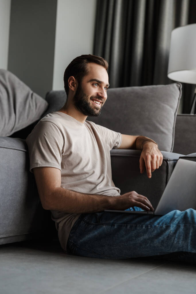 Kép egy mosolygós pozitív fiatalember otthon a kanapén, laptoppal. - Fotó, kép