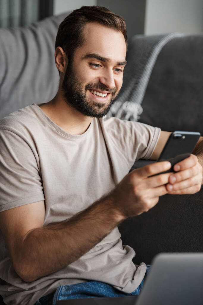 Wizerunek uśmiechniętego, pozytywnego młodzieńca w domu na sofie za pomocą laptopa na czacie przez telefon komórkowy. - Zdjęcie, obraz