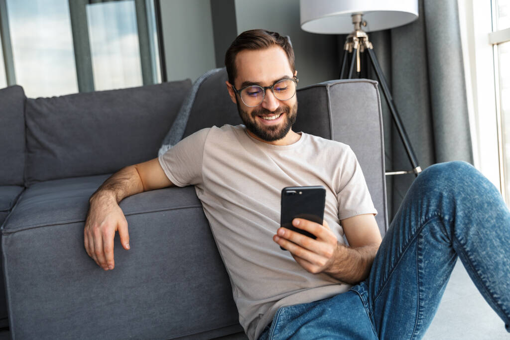 Atractivo joven inteligente sentado en un piso en la sala de estar, utilizando el teléfono móvil
 - Foto, imagen