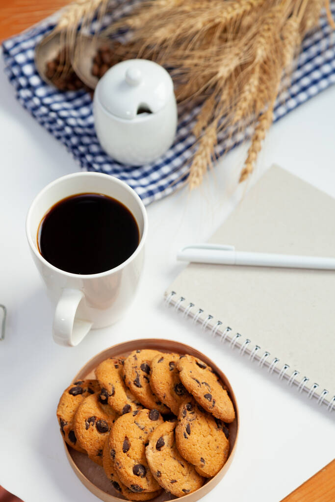 Λευκό φλιτζάνι καφέ και σπιτικά στρογγυλά μπισκότα με κομματάκια σοκολάτας. Το πρωινό είναι στο τραπέζι. Φαγητό στο σπίτι. Νεκρή ζωή. Καφές και κόκκους καφέ. Ένα αναζωογονητικό ποτό. - Φωτογραφία, εικόνα