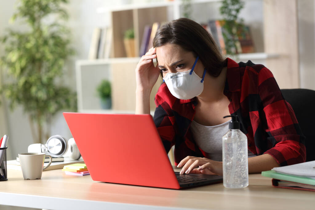 Estudante triste com máscara protetora lendo más notícias coronavírus no laptop sentado em uma mesa em casa
 - Foto, Imagem