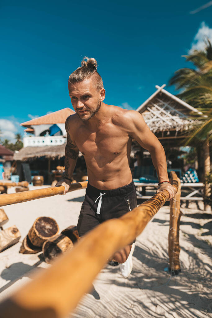 Musculoso hombre de entrenamiento en eco gimnasio en la playa. Equipamiento de fitness de madera exterior
 - Foto, imagen